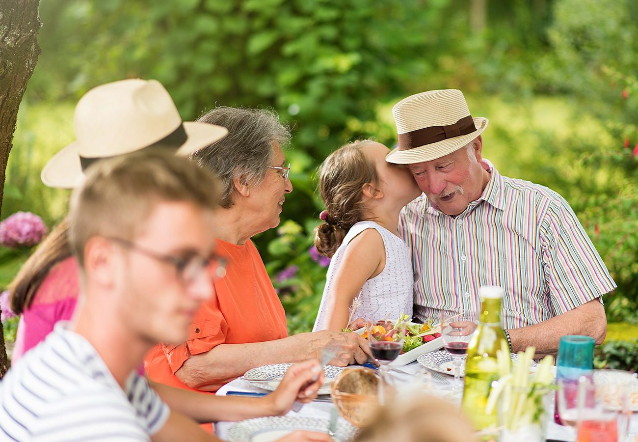A young girl kisses an elderly man on the cheek at an outdoor family gathering, with other people sitting around a table set with food and drinks in a garden.