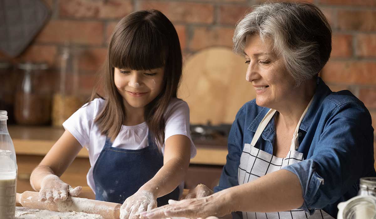 A smiling elderly woman and a young girl, both wearing aprons, roll out dough together on a floured surface in a cozy kitchen. The girl looks happy and focused, enjoying the baking activity.