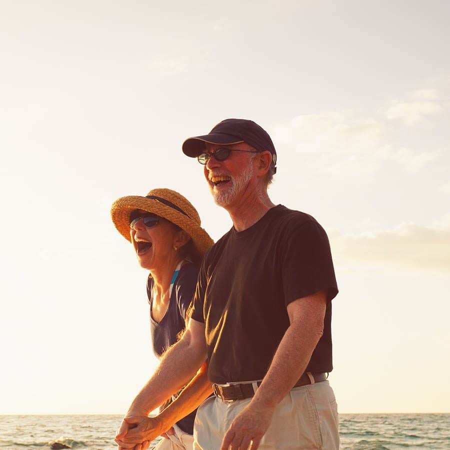 An older couple wearing hats and sunglasses laughs together while walking on a sunny beach, with the ocean and sky in the background.