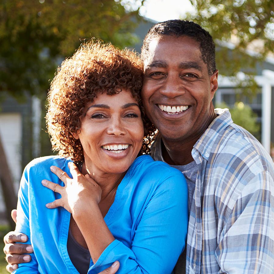 A smiling couple stands close together outdoors, embracing and looking at the camera. The woman has curly hair and wears a blue top, while the man wears a plaid shirt. Trees and a house are blurred in the background.