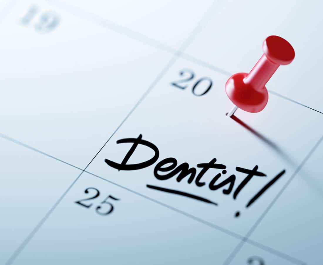 A close-up of a calendar with a red pushpin marking the 20th day. The word Dentist! is written in bold, black letters across that date, indicating a dental appointment.